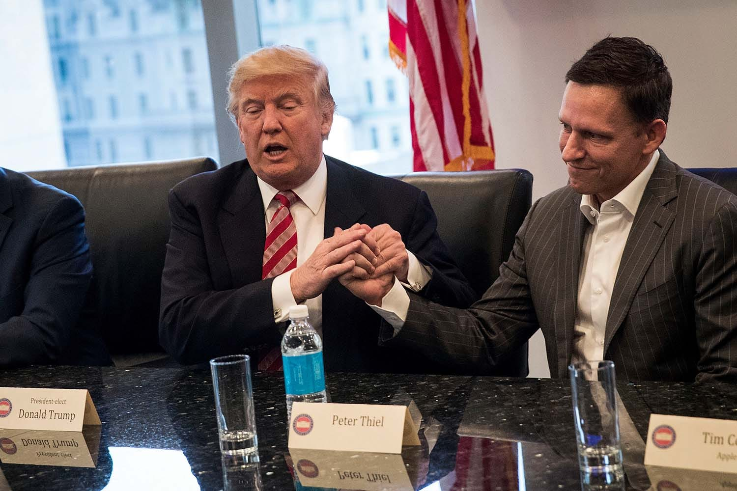 President Donald Trump holding hands with Peter Thiel while seated at a boardroom table.