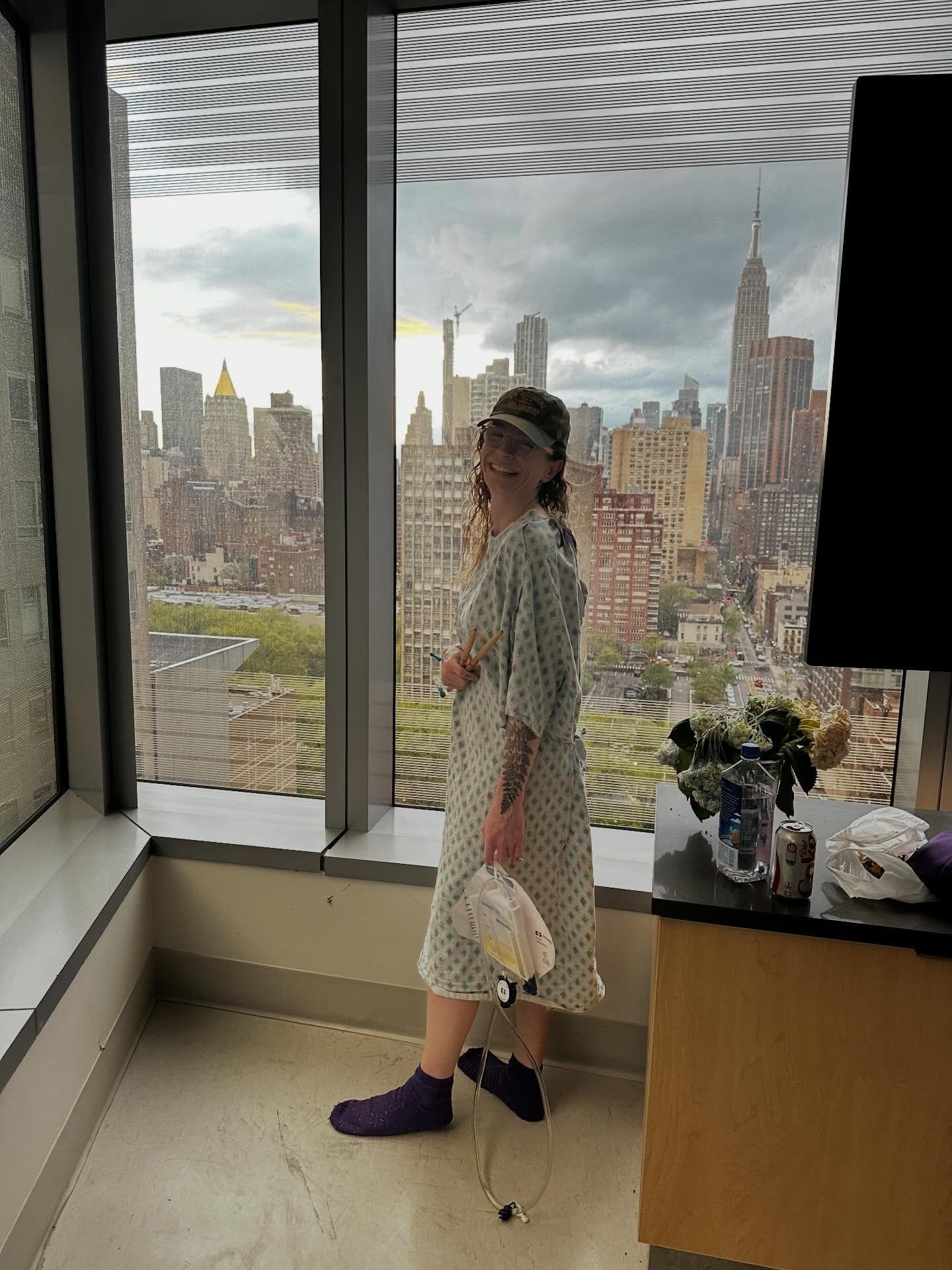 Grace standing in a hospital gown beside the window overlooking New York.