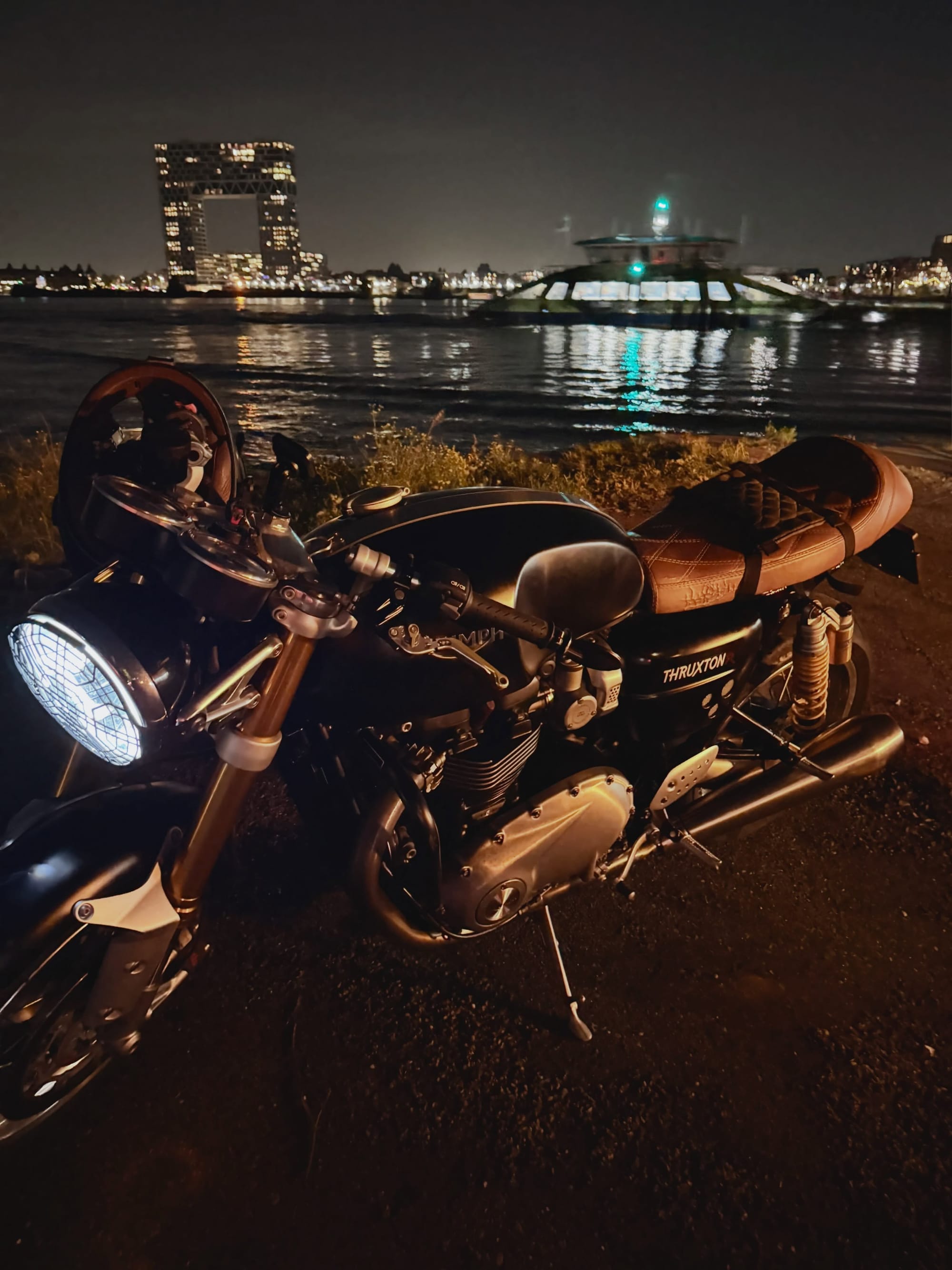 The Triumph parked by the water at night, its headlight illuminated.